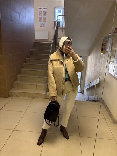 A person is standing on a tiled floor next to a staircase in what appears to be a public building. The staircase has metal railings. A sign is visible on the wall near the stairs in Russian. The person is wearing a beige hat, a beige jacket, light-colored pants, and brown boots. They are holding a phone in their right hand, seemingly taking a photo or video. A black bag is hanging from their shoulder. The scene is illuminated by artificial light. The reflection of the person and the surroundings can be seen on a mirrored surface above. Based on the Russian sign, the location may be in or near Kudrovo, Russia.
