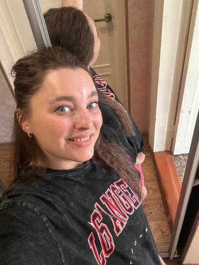 A young woman with light blue eyes and auburn hair is taking a selfie in a room with light-colored walls and wood-paneled flooring. She is wearing a dark grey t-shirt with red and white lettering that appears to say "DREAM BIG". The woman is smiling, and her hair is loose and wavy. The reflection of the woman is visible in a mirror to her left. The room appears to be well-lit, possibly with natural light, though the time of day is not definitively clear. The overall impression is one of casualness and self-expression.