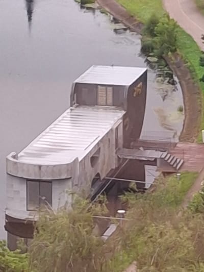 This is a low-resolution, rotated image of a modern, angular building situated on the edge of a body of water. The building has a predominantly white facade with some dark brown elements and large windows. It appears to be a boathouse or a waterside pavilion, with a ramp or walkway leading to its entrance from a green, grassy area. The water in front of the building is calm and reflects the structures.
To the left of the building, a paved pathway curves through a lush green landscape. To the right and behind the building, there is dense foliage, possibly trees or bushes, suggesting a natural or park-like setting. The lighting suggests it is daytime, with no strong shadows indicating a specific time. The overall scene appears peaceful and serene.
There is no visible text in the image.