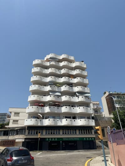 This is a straight-on, medium-angle shot of a distinctive white apartment building with wavy balconies, located in Palma de Mallorca, Spain. The building is tall, with multiple floors, and features numerous rounded openings on each balcony. Several of the balconies have umbrellas and awnings. In the foreground, a grey car is parked on the right side of the frame, with its license plate visible. A traffic light and a street sign are also present. The background shows a clear blue sky and a portion of another building to the right. The overall scene depicts an urban environment on a sunny day.
