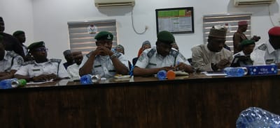 The image depicts a meeting or conference taking place indoors. Several people are seated at a long table with a dark wooden facade. Many of the individuals are wearing uniforms with green berets and shoulder boards that suggest a regulatory, security, or paramilitary service, and some have name tags. Other attendees are dressed in civilian attire, including traditional Nigerian clothing, such as hats and gowns.
Objects on the table include water bottles, drink cans, papers, notebooks, and writing instruments. There's also a box of tissues with a "Nora Happy New Year" design. In the background, there are windows with blinds, air conditioning units mounted on the wall, and a framed poster or chart.
The location is identified as Tudun Wada, Nigeria. The overall setting appears to be an office or meeting room within a government or institutional building.