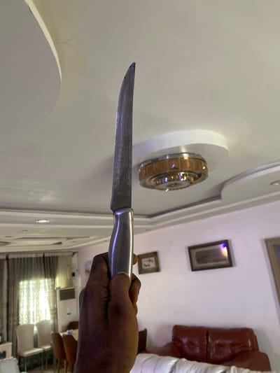 The image shows a person's hand holding a knife upwards, with the blade facing away from the viewer. The background appears to be a residential interior, possibly in Giginyu, Nigeria. The ceiling has ornate light fixtures and a textured design. There are framed pictures on the wall and furniture, including a red leather sofa, visible in the background. The lighting suggests it is daytime, with natural light coming through a window covered by curtains. The overall scene is mundane, with no immediate indication of violence or distress.