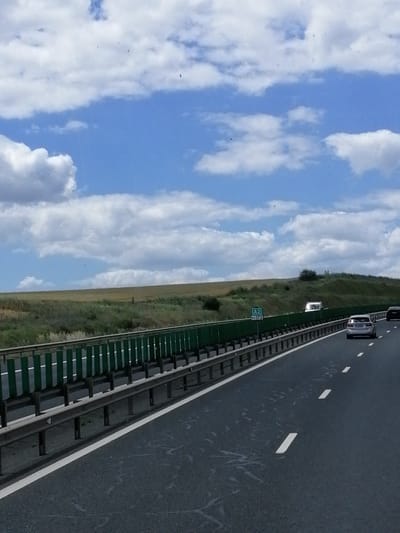 The image captures a view from a car traveling on a highway, likely the A2 in Romania, as indicated by a road sign visible in the distance. The highway features two lanes of asphalt, separated by white dashed lines. On the left side of the highway, a protective barrier with green vertical slats is installed, separating the road from a grassy embankment. Beyond the embankment, rolling hills covered in dry grass and sparse vegetation are visible under a bright blue sky dotted with fluffy white clouds. A few small dark specks, possibly birds or debris, are scattered across the sky. Several vehicles are present on the highway, including a silver car in the foreground and another car further ahead, along with a white camper van parked or moving slowly on the right side. The scene depicts a typical highway travel experience in a rural or semi-rural Romanian landscape on a clear day.