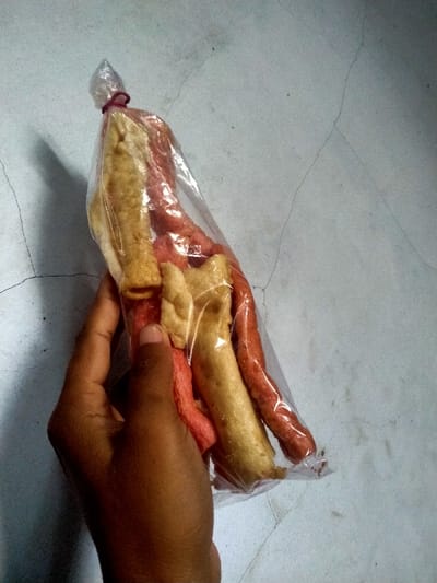 A hand holds a transparent plastic bag containing three long, thin, fried snacks. The snacks are a light yellow and reddish color, and have a slightly irregular shape. One snack is bent in the middle, and the other two are straight. The snacks are in a clear plastic bag with a red rubber band at the top. The background is a white surface. The scene is taking place in a residential area in Kecamatan Sambirejo, Indonesia, based on the coordinates provided.