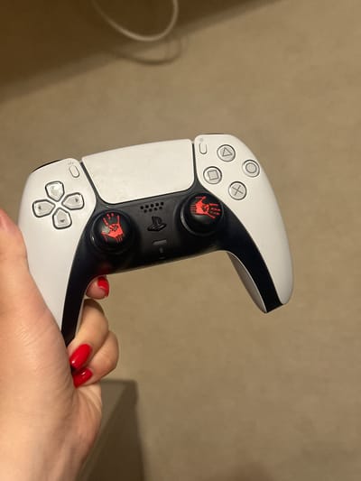 An individual's hand, adorned with bright red nail polish, is holding a white and black PlayStation 5 controller. The controller features custom red thumbstick caps with a stylized hand print design. The background is a plain, light brown surface, likely a carpet or fabric. There are no discernible landmarks or context to suggest a specific location within Lytkarino, Russia.
