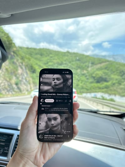 A person's hand holds a smartphone displaying a YouTube video interface, with the video showing a black and white portrait of a woman. The video is titled "Feeling Good Mix - Emma Peters..." and has "57M views" and "5 years ago" beneath it. A "Subscribe" button and "534K likes" are also visible. Below the video are comments, including one that reads "To the 1% who's reading this, may your dreams come true, stay safe and have a wonderful day..." Another video titled "OMER BALIK Mix - Best of 2021 [2 Hours]" is partially visible at the bottom of the screen. The phone is held in front of the viewer in a car interior, with a blurry view of a road and green, mountainous landscape seen through the windshield. The location is identified as Stanišići, Montenegro.