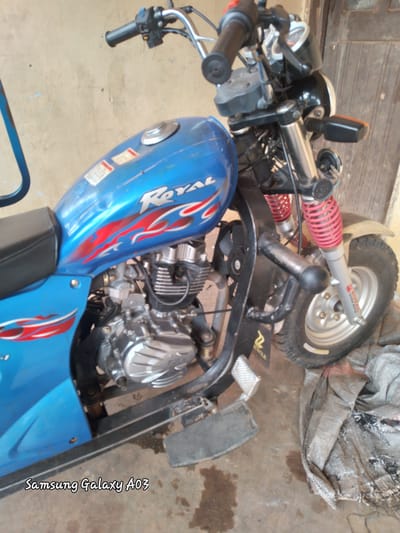 The image shows a blue motorcycle with red and white flame graphics parked on a dirt ground in front of a textured, light-colored wall. The motorcycle has a black seat, a silver engine, and red shock absorbers. A wooden door is visible on the right side of the image. The lower right corner shows a torn grey burlap sack. The text "Samsung Galaxy A03" is superimposed in the lower left corner of the image. The scene is located in Dokan Tofa, Nigeria.
