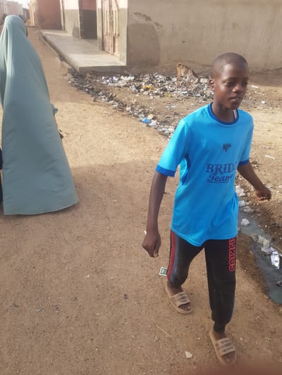 The image shows a boy standing on a dirt street in Dutsinma B, Nigeria. He is wearing a blue t-shirt with "Bride Team" printed on it, black pants, and sandals. There is trash visible in the background. A person wearing a light green robe is partially visible in the foreground. The street is unpaved and dusty. A building is visible to the left.