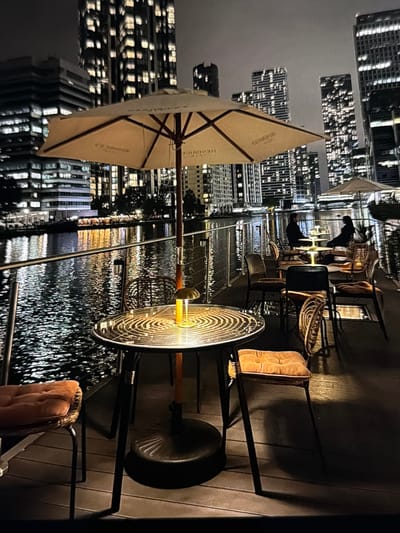 The image depicts a nighttime outdoor patio located beside a body of water in the city of Maidstone, United Kingdom. The foreground features a large, light-colored patio umbrella bearing the "HENDRICK'S" brand name. Beneath the umbrella is a round table with a dark, patterned surface, illuminated by a small, glowing yellow table lamp. Adjacent to the table, a chair with an orange cushioned seat and wicker-like back is partially visible. The immediate ground consists of dark wooden decking. A metal railing separates the patio from the water.
In the midground, the body of water reflects numerous bright city lights, creating shimmering patterns. Further down the patio, several other tables with similar lamps and chairs are arranged, with two individuals seated at one table, appearing to be in conversation or dining. A smaller, light-colored umbrella is also visible in the distance. The background is dominated by a city skyline comprising multiple high-rise buildings with numerous illuminated windows, indicative of an urban environment at night. The sky is dark and overcast.