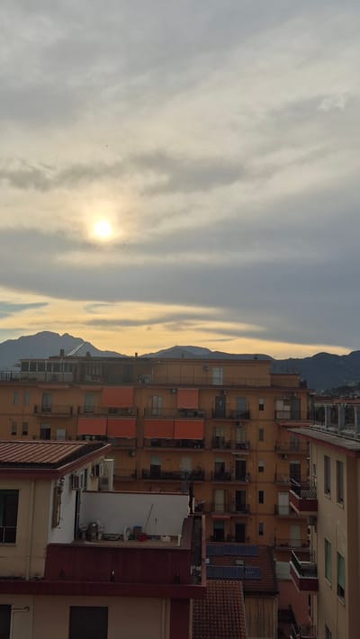 The image captures a view of Salerno, Italy, on a cloudy day, likely during sunset or sunrise. The foreground features the rooftops and upper floors of several buildings, constructed with a mix of light and dark red tile roofing, alongside light-colored stucco and orange-painted facades. One building in the mid-ground exhibits balconies with orange awnings and numerous windows, suggesting residential apartments. Another building to the right has a similar architectural style with balconies and windows. In the background, a range of mountains is visible under a sky characterized by grey and yellowish clouds, with the sun partially obscured, creating a diffused light. The scene appears to be an urban residential area, with a focus on the architecture of the buildings and the natural backdrop of the mountains. There are no visible people in the image, and the overall impression is that of a quiet, urban landscape.