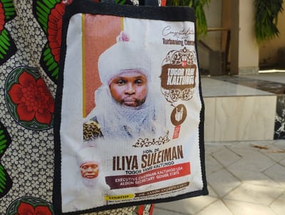 The image shows a white tote bag with black handles. The bag is printed with a picture of a man wearing a white turban and white robe.  The man is looking directly at the camera and has a serious expression on his face. The text on the bag reads "Congratulations on your Turbaning Ceremony", "TOGOR YAM KALTUNGO", "HON. DR. ILIYA SULEIMAN TOGOR YAM KALTUNGO", "EXECUTIVE CHAIRMAN KALTUNGO LGA ALGON SECRETARY GOMBE STATE", and "COURTESY MR. AARON ANDREW LABTE". The bag is being held by a person wearing a floral dress. The image was taken in Kaltungo, Nigeria, at the coordinates (latitude: 9.816, longitude: 11.315). The image is taken from an angle that shows only the front of the bag. The background of the image is a blurry view of a tiled floor.  There is a small amount of green foliage visible in the top right corner of the image.  The image is well-lit.