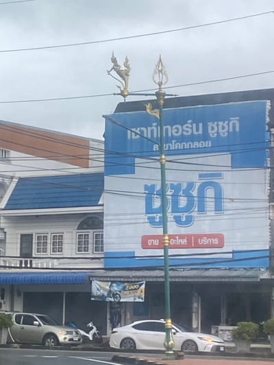 The image depicts an outdoor commercial street scene in Khok Kloi, Thailand, viewed under an overcast sky during the daytime. The prominent feature is a large blue billboard occupying the upper-right portion of the frame. This billboard advertises "Southern Suzuki" (เซาท์เทอร์น ซูซูกิ) with a specific mention of its "Khok Kloi branch" (สาขาโคกกลอย). The word "Suzuki" (ซูซูกิ) is displayed in large white letters, and a red rectangular section specifies "Sale | Parts | Service" (ขาย อะไหล่ | บริการ).

In the foreground, a tall green street light pole is adorned with ornate gold decorations. These include a golden, mythical creature resembling a garuda near the top-left, and a trident-like finial at its apex. Numerous dark utility cables crisscross the sky, extending across the frame.

Along the right side of the street, several vehicles are parked, including a white sedan, a silver pickup truck, and a white scooter. Buildings with blue awnings or roofs and white facades featuring multi-paned windows line the street. A smaller yellow banner, partially visible below the main billboard, displays the number "7,000". No people are visible in the scene, which captures a typical daytime commercial environment in the specified location.