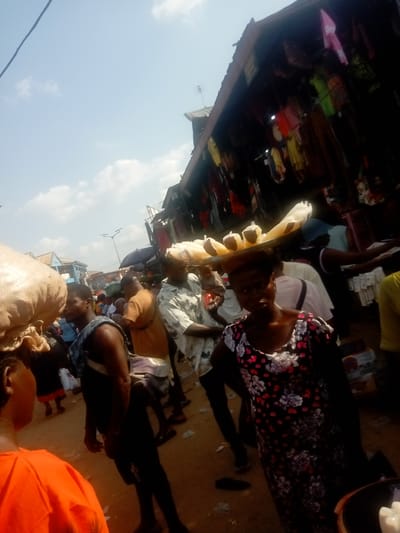 The image shows a busy street scene in Awka, Nigeria. The scene appears to be a marketplace. People are gathered closely together, walking in different directions. A woman in a floral dress is carrying a tray of what appears to be food items on her head. Clothes are hanging from a building in the background. The ground is dirt. The sky is blue with light clouds.