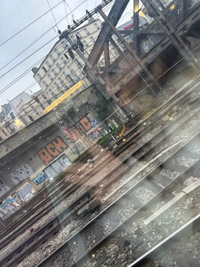 The image depicts an urban railway environment in Bobigny, France, viewed from an elevated perspective under an overcast sky. Multiple parallel railway tracks, comprising steel rails, sleepers, and ballast, diagonally traverse the lower and mid-sections of the frame. Overhead electrical lines and their support structures, including poles and gantries, are visible above the tracks. In the midground, a concrete retaining wall is extensively covered with various graffiti tags, notably large orange "ACH" lettering and blue "nes" tags. Behind this wall, a dark metal truss bridge or viaduct structure, featuring a metal fence, is present. In the background, several multi-story buildings with light-colored facades and numerous windows are visible, one displaying a partial red and white sign reading "HOND." Yellow safety markers or equipment are positioned near the tracks in the mid-distance. No individuals or specific actions are depicted in the scene.