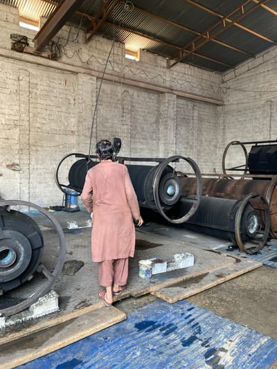This image depicts an indoor industrial environment within Daman, Afghanistan. A single adult male, viewed from behind, wearing a light brown shalwar kameez and sandals, is walking towards the right side of the frame. He has dark hair. The scene is dominated by large, horizontal, cylindrical industrial machinery, likely rollers, painted black with circular end plates, some featuring a central blue component. These rollers are supported by metal frames and appear to be arranged in a series along the right side of the space. Another similar roller is partially visible on the far left. The floor consists of concrete, partially covered by blue corrugated plastic sheets and several flat wooden planks, with visible wet areas or puddles. Near the man's feet, on the floor, are a white bucket with paint stains and a grey rectangular block. The walls are constructed from rough-textured, light-colored brick. The ceiling is a corrugated metal roof supported by metal beams, with natural light entering through several rectangular openings. An overhead crane or hoist system, including a motor, wires, and a hanging hook and cable, is visible near the ceiling.