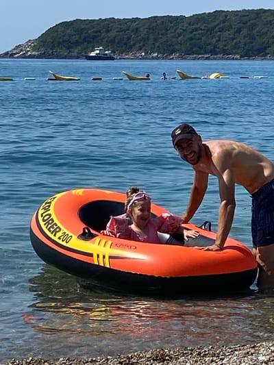 A man and a young girl are in an orange and black inflatable raft on the water. The man is shirtless and wearing dark blue shorts, leaning over the girl. The girl is wearing a pink shirt and has a purple headband on. The raft has "EXPLORER 200" written on it. The water is a clear blue, with some ripples. In the background, there is a lush green hillside with some rocky outcrops. Several yellow buoys are visible in the water, marking a boundary. The sky is a clear blue, suggesting a sunny day. The scene appears to be a recreational activity at a beach or lake. The man seems to be smiling and engaged with the girl. The overall mood is lighthearted and summery. The location is Prijevor, Montenegro.