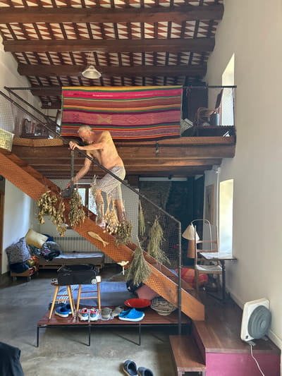 This media file captures an indoor scene with a man and various objects, possibly in a home or workshop. The man, appearing to be of older age, is shirtless and wearing plaid shorts. He is in a contorted pose, leaning on a railing of a wooden staircase that is adorned with dried plants and a fish-shaped decoration. He seems to be in the process of navigating the stairs.

The setting is characterized by rustic wooden elements, including pillars and the staircase itself, which create a sense of a unique, possibly custom-built interior. A colorful, striped rug hangs on a wall made of wooden planks with triangular patterns. Other objects visible include a ceiling fan, a lantern, a shoe rack with several pairs of shoes, pillows, and bags. The lighting suggests it's daytime, with natural light filtering in from above. The overall impression is one of a lived-in, eclectic space with a focus on natural materials and a touch of bohemian decor. There is no visible text in the image. The location context of Pineda de Mar, Spain, is provided externally.