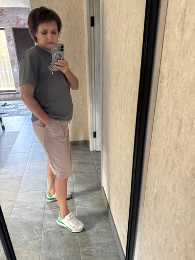 The image shows a woman standing in a room, likely a house, taking a mirror selfie. She is wearing a grey t-shirt and light pink shorts. She has her right hand in her pocket. She is wearing white sneakers with green accents. The room has tiled floors and beige textured walls. In the background, a door and a window with blinds are visible. The woman's hair is dark and styled in a bob. The reflection shows a door and a brick wall with a window. The overall scene suggests a casual indoor setting.