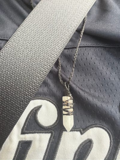 The image shows a close-up of a necklace with a crystal pendant resting on dark fabric with white lettering. A grey, textured strap is diagonally positioned across the upper left portion of the frame. The fabric beneath the necklace appears to be part of a garment, possibly a jersey, due to the patterned texture and the prominent white lettering, which may be an initial or logo. The crystal pendant is elongated and multifaceted, wrapped with dark wire. The scene is devoid of any recognizable urban or natural features that would indicate a specific location in Vallensbæk Landsby, Denmark. The lighting suggests an indoor setting or an overcast day.