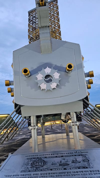Here's a detailed description of the image:
**General Description:**
The image showcases a portion of an art installation or public artwork, likely situated outdoors. The structure appears to be a stylized representation of a Korean turtle ship (Geobukseon), a type of armored warship historically used by the Korean navy. The photo is taken at an upward angle, looking towards the sky. The orientation is somewhat unusual, making it appear as if the structure is floating.
**Specific Elements:**
* **Turtle Ship Representation:** The main focus is a grey, boxy structure with several golden cylindrical protrusions. A prominent design element is a circular emblem on the front, containing a yin-yang symbol at the center surrounded by white leaf-like patterns. A metal lattice structure extends from the side, representing parts of the ship.
* **Signage/Plaque:** Adjacent to the ship structure is a large sign or plaque bearing text in Korean and what appears to be an image of an old Korean banknote. The text on the sign makes references to the HD Hyundai Heavy Industries ship building tradition and a 500 Won note.
* **Environment:** The structure is enclosed by metal railings and fencing. The background features a blurred view of what appears to be an urban landscape.
**Location:**
Based on the presence of references to HD Hyundai Heavy Industries in the signage, this structure is likely located in the city of Ulsan, South Korea, a major shipbuilding center and the location of HD Hyundai's main operations.
**Overall Impression:**
The image depicts a commemorative artwork celebrating Korean shipbuilding history. The unconventional framing and viewpoint add an element of visual interest.