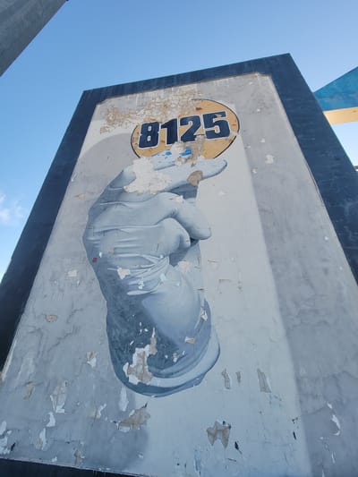 The image shows a close-up of a weathered sign with a graphic of a hand wearing a glove, painted in shades of gray and white. The hand is holding a circular emblem with the number "B125" in dark blue lettering against a yellow background. The sign's surface is heavily cracked and peeling, with patches of paint missing, revealing the underlying material. The sign appears to be mounted on a larger structure, with a dark blue border along its top edge. Above the sign, a bright blue sky is visible, suggesting it is daytime. The sunlight casts shadows and highlights on the sign, emphasizing its texture and age. The overall impression is one of decay and wear, with the vibrant graphic of the hand contrasting with the degraded state of the sign. The image's perspective is tilted, giving a dynamic and somewhat abstract view of the sign. No specific location cues beyond the general context of a sign are evident in the image itself. The weather appears to be clear and sunny.