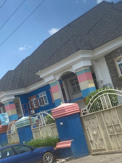 The image depicts a colorful building with a dark, multi-peaked roof, situated in Iddo Tudunwada, Nigeria. The building's exterior walls are painted a vibrant blue, accented with horizontal bands of pink, yellow, and light blue on decorative pillars and sections of the facade. White trim frames the windows and roofline. Several windows, some open, are visible, featuring white frames and grilles. A large, ornate metal gate, cream-colored and decorated with intricate scrollwork and geometric patterns, stands in front of the building, partially obscuring a white metal fence with decorative elements. Green bushes are visible behind the fence and gate. A blue sedan is parked in the foreground, angled towards the right side of the frame. Power lines are visible diagonally across the bright blue sky, which has a few scattered white clouds. A blue banner with white text is attached to the left side of the building. The scene is captured in daylight, with the sun casting shadows that are not clearly discernible.