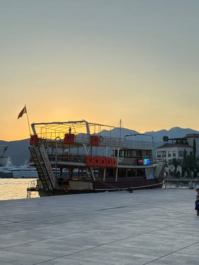 A sunset scene unfolds in Tivat, Montenegro, where a large, multi-decked tour boat, painted in shades of brown and cream, is docked at a paved promenade. The boat features several life preservers attached to its exterior. A small red flag flies from the top deck. In the background, other yachts and buildings, including a large white luxury yacht, are visible. The sky is a gradient of soft orange and pale blue, with the sun setting behind distant mountains. Two young children stand on the promenade in the foreground, one wearing a blue shirt and the other a pink shirt. The water is calm and reflects the warm light of the sunset.