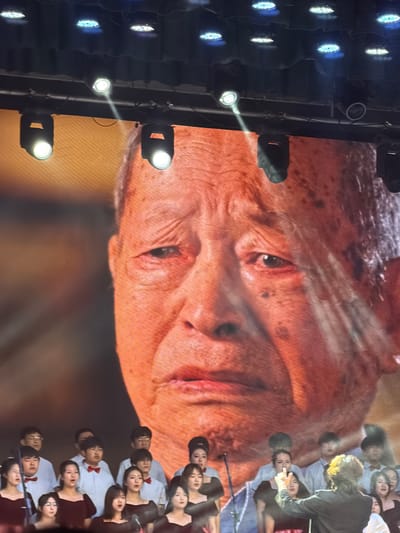 This image captures a vibrant performance scene, likely indoors at a concert hall or auditorium in Guta, China. The primary focus is a massive, close-up projection of an older person's face on a large screen, occupying the upper and central parts of the frame. The face displays deep wrinkles and a reddish-orange skin tone, possibly due to screen display or stage lighting, conveying a weathered and contemplative expression.
On the right side of the screen, partially in front of the projected face, a group of people is visible. Many are wearing white shirts, and some sport red bow ties, strongly suggesting they are a choir or a performing group. Their faces are turned towards the left, with some appearing to sing or react to the performance, and others looking attentively forward.
To the far left of the image, a vertical array of bright stage lights illuminates the scene. Several lights emit strong, concentrated beams across the space, highlighting the performance area. The overall lighting setup indicates an indoor evening event. No discernible text is visible in the frame. The combination of the large projected face, live performers, and professional stage lighting points to a significant public event, possibly a musical performance, concert, or theatrical production.