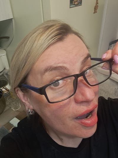 A fair-skinned female is depicted in a medium close-up, high-angle photograph, likely within a domestic setting in Elektrostal, Russia. She has blonde hair and is wearing a black top with a thin gold-colored chain. Her left eye (viewer's right) is wide open and directed towards the camera, while her right eye (viewer's left) is partially obscured by a pair of eyeglasses. She is holding the black-framed eyeglasses, which feature blue temples, with her right hand; her fingernails are painted light pink. Her mouth is slightly agape, revealing teeth and tongue, with lips colored in a peachy-pink lipstick. A silver-colored hoop earring with dark stones is visible on her left ear. The background consists of light-colored walls. To the left, a white fixture (possibly a lampshade), a clear glass, and a patterned surface are visible. To the right, a framed picture depicting a partially nude figure and a dangling decorative item are mounted on the wall.