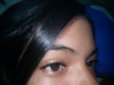 The image is a close-up of a person's right eye and forehead, taken from a slightly elevated angle. The subject has dark hair that partially obscures the left side of the frame. The eye is open, with dark brown iris and eyelashes. The skin appears to have a light brown complexion. The background is dark and out of focus, with a blue fabric visible on the right side of the image. There is no indication of the city of El Maco, Venezuela, or any specific location within it.
