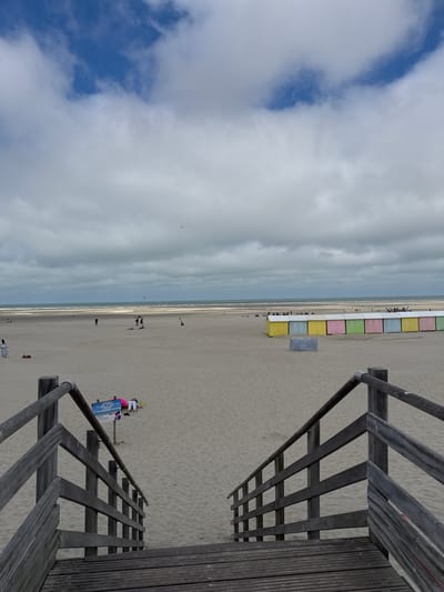 The image depicts a wide, sandy beach in Berck, France, under a cloudy sky. In the foreground, a wooden staircase leads down to the beach. Further down the beach, a row of colorful beach huts, painted in yellow, blue, pink, and green, are visible. Several people are scattered across the sand, some walking and others relaxing. The ocean stretches out in the background, with faint waves and a distant horizon. There are also some kites visible in the air, suggesting a windy day. A sign on the beach appears to be a "Plage sans déchets" (trash-free beach) sign.