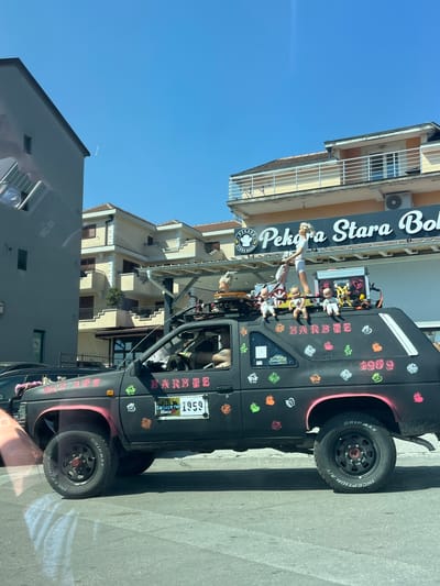 The image shows a black modified truck parked on a street in Mrčevac, Montenegro. The truck is decorated with various stickers, including pink heart shapes and cartoonish figures. Several dolls are placed on the roof of the truck, some appearing to be in playful poses. The words "BARBEE" and "GARBAGE RUN ALBANIA 2026" are visible on the side of the truck, suggesting it might be part of an event or a custom build. A license plate reads "1959".
In the background, there is a multi-story building with balconies and an air conditioning unit, and a sign that says "PEKARA STARA BOKA" which likely indicates a bakery. The sky is clear and blue, suggesting a sunny day. The scene appears to be a street in a town or city, with buildings and a paved road. The time displayed on a digital clock in the foreground is 12:38. The overall impression is a bright, possibly festive, street scene with a uniquely decorated vehicle.