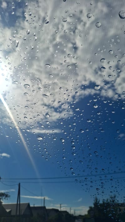 The image shows a close-up view of water droplets on a glass surface, likely a car window, with a bright blue sky and white clouds in the background. The droplets are of various sizes, some appearing spherical and others more elongated, reflecting the light and the sky. A sunbeam cuts diagonally across the lower right portion of the frame, creating a lens flare effect. On the far right, a dark, blurred silhouette suggests trees and buildings, with power lines visible against the sky. The presence of raindrops indicates recent or ongoing precipitation, suggesting a cloudy or rainy weather condition, despite the bright blue sky visible through the window. The image seems to capture a moment after rain, with the sun beginning to break through. The scene evokes a sense of peacefulness or contemplation, often associated with watching rain from indoors or from a vehicle. There is no visible text in the image.