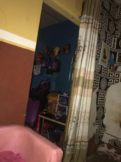 The scene depicts an interior space, likely residential, located in Oba-Ile, Nigeria. The perspective is from one room looking into an adjacent, smaller room through an open doorway, with another section of the originating room visible on the right.
In the foreground, to the lower left, a portion of a pink upholstered armchair or sofa is visible, partially covered by pink fabric. The wall immediately behind this furniture piece features a horizontal division: a reddish-brown lower section, a thin white stripe, and a yellowish-orange upper section.
The open doorway leads into a room with solid blue-painted walls. Hanging on the blue wall are several items: three caps or hats (one yellow, one red, one striped), a framed photograph, and at least four bags of varying designs (including white with black patterns, black with pink patterns, and blue). On the floor or stacked within this inner room are a tall, purple, possibly luggage-style container; a large, orange, perforated laundry basket; and a smaller, multi-tiered purple shelving unit holding various small objects. A dark-colored table or counter is partially visible, with a dark blue bucket situated on the floor beneath it.
To the right of the doorway, a striped, floor-length curtain with a beige, white, and brown ornate pattern is pulled aside. It partially obscures a wall covered in black and white geometric pattern wallpaper. On this patterned wall, a white-framed picture with a yellow background features a dark silhouette of a person and the text "ALHAMDULILLAH". Below this frame, several small items resembling key holders or decorative elements are attached to the wall. The lower portion of this wall displays an irregular, lighter-colored patch, suggesting damage, peeling, or an unfinished section over a reddish-brown base. No individuals are present in the image.