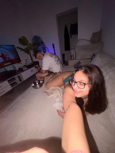 This image depicts an indoor scene, likely a living room or bedroom, where two individuals are present. A man, appearing to be in his early twenties, is seated on the floor, looking at his phone. He is wearing a white t-shirt with graphics on it and dark shorts. Beside him, a metal bowl rests on the floor. In the background, a television screen displays a game or show, and a potted plant with large leaves is visible. To the right of the man, a woman is lying down, facing away from the camera and towards the man. She is wearing teal colored bottoms and is partially covered by a blanket. Her arm is extended towards the camera, obscuring part of the foreground. The room is dimly lit, with artificial lighting contributing to the overall atmosphere. The presence of a Stitch balloon suggests a playful or relaxed setting. The location of Mönchengladbach, Germany, is not discernible from the visual elements of the image.