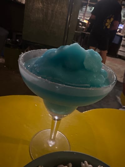 An illuminated, frozen, blue cocktail sits in a margarita glass with a salted rim, resting on a bright yellow table. In the background, a person wearing a black t-shirt with a yellow flower design is visible, standing in a dimly lit environment. The scene suggests a relaxed outdoor or indoor setting, potentially a bar or restaurant. No specific location in Larnaca, Cyprus can be determined from the image.