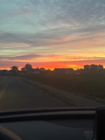 The image depicts a sunset scene with an orange and red-streaked sky over what appears to be a road and residential area. The right side of the image shows the interior of a vehicle, possibly a car. Trees and buildings are silhouetted against the bright sky, suggesting the presence of a town or city. Based on the prompt, the location is Tourcoing, France, but specific identifying features beyond the general urban/suburban environment are not discernible from the image.