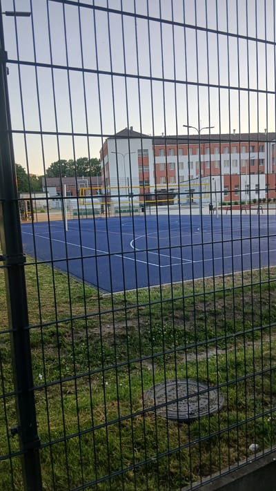 The image shows an outdoor sports court with a blue surface, marked for volleyball and basketball. A volleyball net is set up across the court. In the background, a modern residential building with white and orange-red facades is visible. Trees and other smaller buildings are also present. The foreground is dominated by a black metal fence, with green grass and a manhole cover in front of it. The scene is set in the city of Jelgava, Latvia, likely in a residential area or a school complex. The sky is clear and light blue, suggesting it's daytime, possibly late afternoon or early evening. No people are present in the frame.