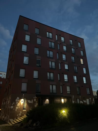 A multi-story apartment building in Vantaa, Finland, stands under a twilight sky filled with dark, cloudy formations. The building's facade is a deep, reddish-brown, segmented by vertical and horizontal lines, with rows of windows and some balconies. The lower portion of the building features a mural with white, stylized floral or leaf patterns. Exterior lighting illuminates an outdoor staircase leading up to the building's entrance, and another light shines on a nearby patch of dense green bushes. The ground in the foreground is paved with dark interlocking stones.