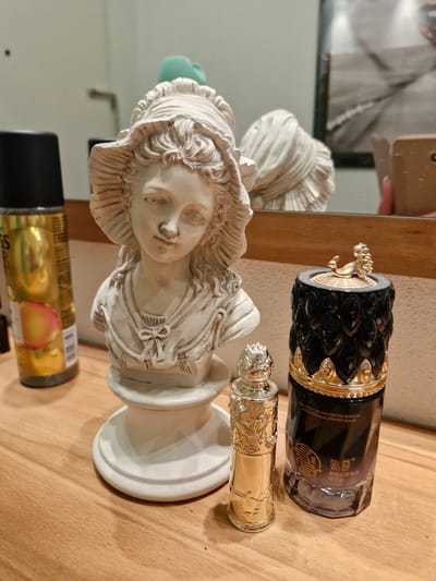 The image depicts a collection of decorative objects and cosmetic bottles arranged on a wooden surface, likely a dresser or shelf. In the foreground, a white bust of a young woman with a wide-brimmed hat stands prominently. To the left of the bust is a tall, metallic gold spray bottle with a black cap. To the right of the bust are two smaller decorative bottles. One is slender and metallic gold, adorned with intricate floral patterns. Adjacent to it is a larger, dark bottle with an ornate black and gold lid featuring a small golden animal sculpture. The background shows a reflection in a mirror, revealing a portion of a room with a framed picture and hints of other objects. There are no people present in the scene. The image provides no information about the city of Bremen, Germany.