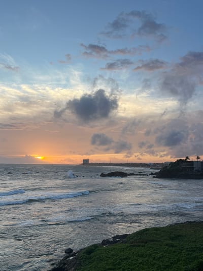 The image depicts a serene coastal scene at sunset in Galle, Sri Lanka. The sun is low on the horizon, casting a warm, golden glow across the sky and the calm ocean. Wispy clouds, tinged with pink and orange, are scattered across a pale blue sky, creating a dramatic and picturesque atmosphere.
In the foreground, the gentle waves of the Indian Ocean lap against a rocky shoreline. The water is a shimmering silver, reflecting the colors of the sunset. To the right, a lush green embankment, likely a manicured lawn or hillside, slopes down towards the water.
Further in the distance, a coastal town is visible, with buildings and palm trees silhouetted against the vibrant sky. The overall mood is one of tranquility and natural beauty, typical of a tropical sunset. The time of day is clearly sunset, indicated by the low sun and warm lighting. There is no visible text in the image.
