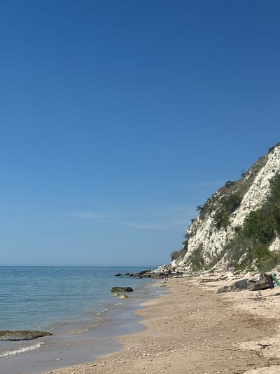 The image depicts a scenic beach in Topola, Bulgaria, under a clear blue sky. The foreground features a sandy beach with scattered rocks and pebbles. Gentle waves lap at the shore, with the water being a calm, translucent blue. To the right, a steep, white chalk cliff rises from the beach, covered in sparse green vegetation. A lone person is walking along the shoreline, away from the viewer, towards the cliff face. Further down the beach, several large rocks and a green pole are visible, along with what appears to be a towel draped over a rock. The overall scene conveys a sense of tranquility and natural beauty characteristic of a seaside location.
