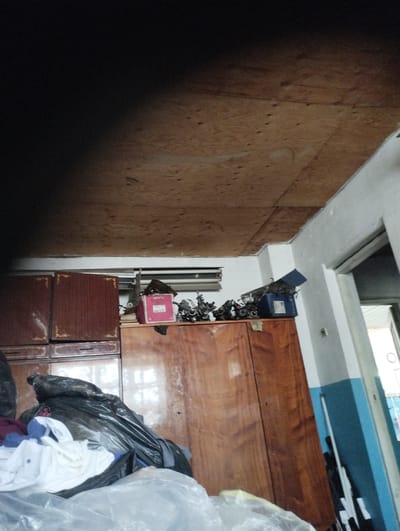 A cluttered room in Tiraspol, Moldova features a wooden cabinet with several items on top, including a pink box and a blue electric device. Above the cabinet, a ceiling made of plywood panels is visible. In the foreground, piles of laundry and plastic bags are stacked. A doorway leading outside is to the right of the image, with a blue and white painted wall next to it.