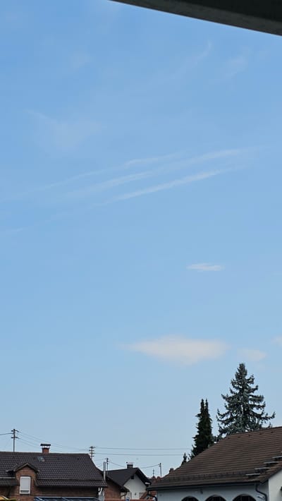 The image shows a view of the sky and rooftops in Vöhringen, Germany. The sky is a clear blue with some wispy white clouds. In the foreground, there are several rooftops of houses, some with chimneys. There are also some trees visible, including a tall pine tree. Power lines and utility poles are also present. The overall scene suggests a residential area in a town or city.