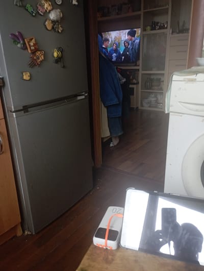 The image depicts an interior scene, likely a domestic living space in Olaine, Latvia. A large, gray refrigerator dominates the left side of the frame, adorned with various magnetic decorations, including cartoonish figures, animals, and what appears to be a small bee. To the right of the refrigerator, a doorway leads to another room where a television is visible, displaying an image of several young males. Next to the television is a shelving unit filled with various items, including what seem to be dishes and decorative objects. In the foreground, a white portable power bank with an orange strap is placed on a wooden surface, alongside a tablet displaying a reflection of the photographer. The flooring appears to be hardwood. The overall impression is of a lived-in, functional space.