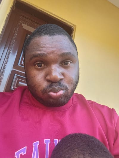 A medium shot depicts a Black man wearing a red shirt with lavender lettering, seemingly a collegiate or team name. He has a trimmed beard and short, dark hair. The background includes a decorated brown door frame and a yellow wall. The location is Suleja, Nigeria.