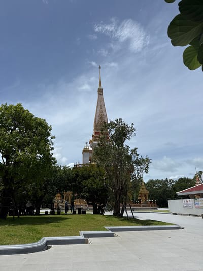 A tall, ornate, cream-colored spire topped with a golden finial dominates the center of the frame, its intricate details visible against a pale blue sky with scattered white clouds. The spire is part of a larger temple complex, indicated by glimpses of gold-trimmed architecture and other decorative elements behind it. In the foreground, a lush green lawn is bordered by a gray, curved concrete walkway. Several trees with dense green foliage are strategically placed, partially obscuring the temple structure. In the middle ground, a few individuals are visible walking on the grass or standing near some benches. To the right, a white building with a red roof is present, along with some construction barriers and signage. The overall scene suggests a well-maintained outdoor area within a temple or religious site in Chalong, Thailand.
