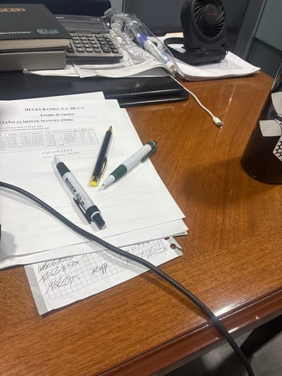 This is a photograph of a wooden desk in an office environment, likely located in Cuautitlán Izcalli, Mexico. The desk is covered with various items, including a black calculator, a stack of papers with financial statements, a black marker, two pens, a small black fan, and a black mug. There are also some handwritten notes on the papers. The overall impression is one of a workspace that is actively being used, with documents and writing instruments laid out. The lighting suggests it is daytime, and the environment appears to be indoors.