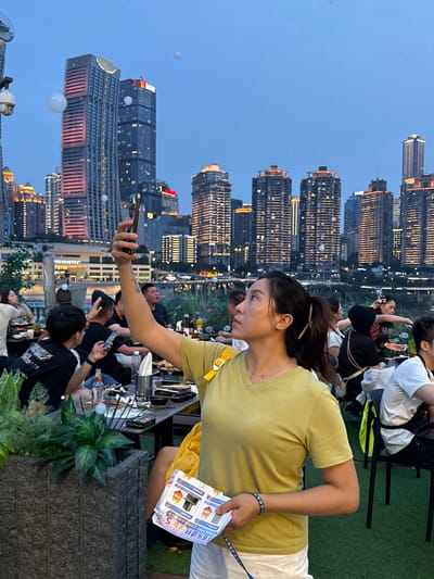 This is a vertical, medium shot of a woman taking a selfie with her phone, while sitting at a table outdoors. She is wearing a lime green t-shirt and white pants, with a blue beaded bracelet on her left wrist. In the background, a city skyline with numerous illuminated high-rise buildings is visible against a dusk sky. Other people are seated at various tables, some holding phones and appearing to take pictures or videos. The scene suggests a lively evening gathering at a restaurant or bar with a scenic city view. The overall atmosphere is casual and social. Visible text in the image includes "WEID CLUB" and promotional text in Chinese, which appears to be an advertisement for "Jiale Jiancai" (多乐建材) offering a discount of 5 units and a "trade-in" promotion for fireproofing materials. The time of day is dusk, with the city lights beginning to dominate the darkening sky. The weather appears to be clear. The setting is an outdoor elevated area, likely a rooftop or balcony, overlooking a river or waterfront.