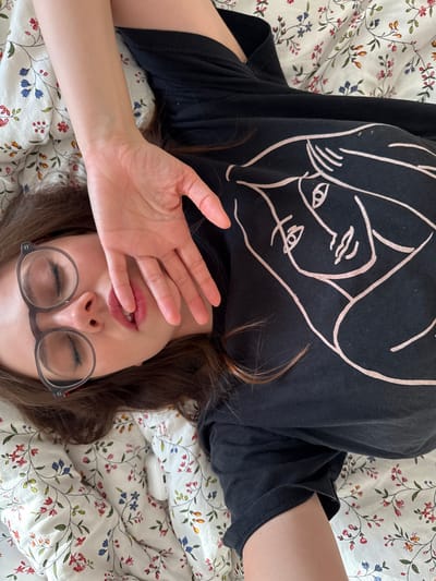 A person with brown hair and glasses lies on a bed with a floral patterned sheet. The person is wearing a black t-shirt with a white line art design of faces. Their left hand is raised, partially covering their face, and their eyes are closed. The scene appears to be indoors, with natural light. The person is the central focus. There is no information to determine the exact location of this image.